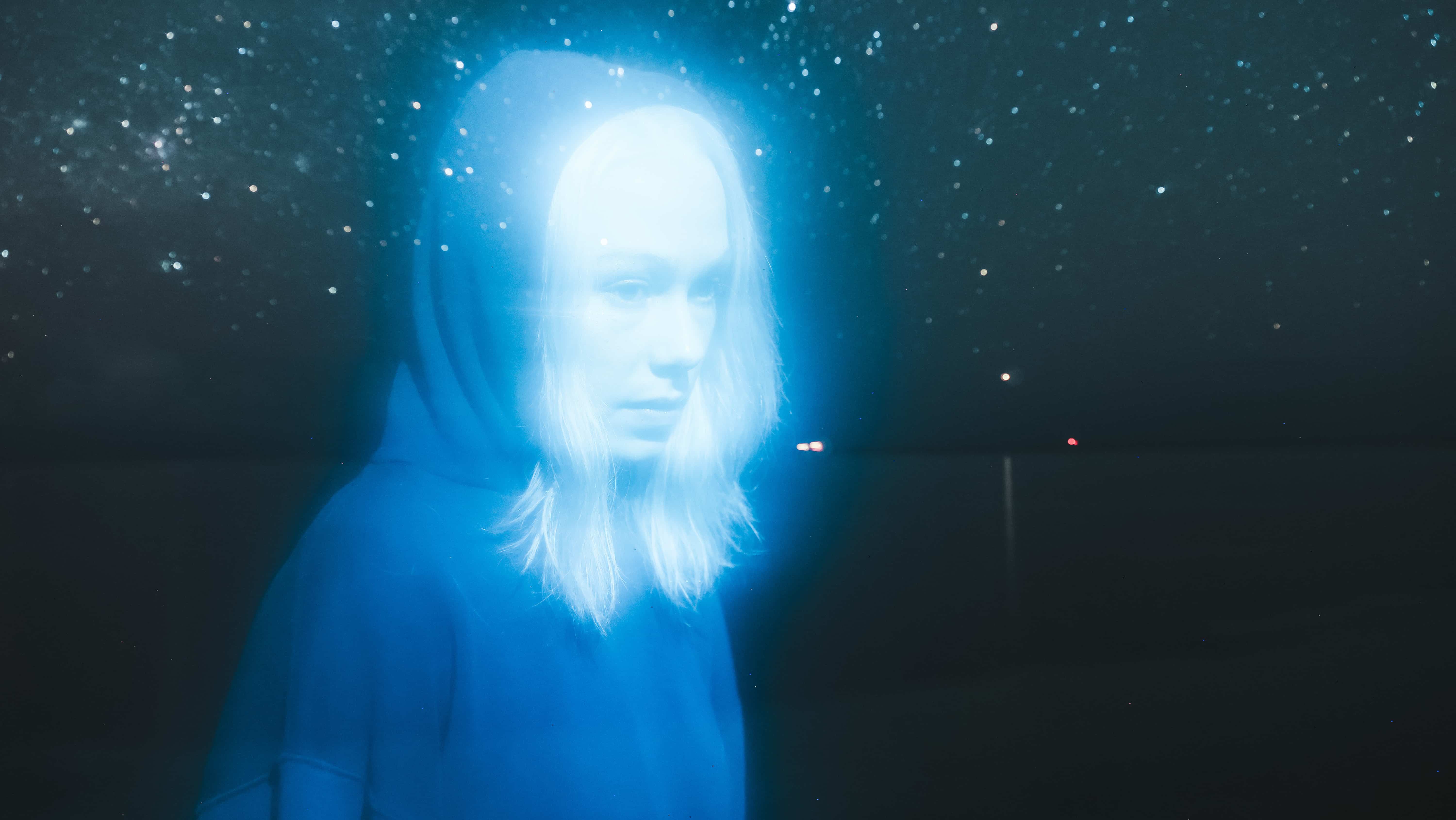Phoebe Bridgers wears a hoodie with a background of stars and space. The image is bathed in blue. Photo: Olof Grind