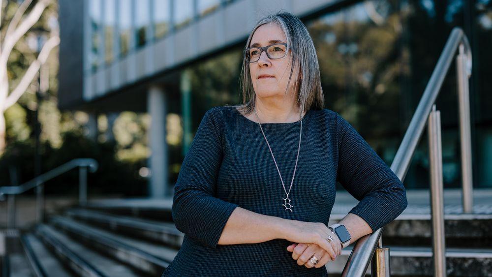 Jacqui Cameron stands thoughtfully near modern building stairs, wearing a dark dress, silver necklace, and smartwatch, in outdoor daylight.