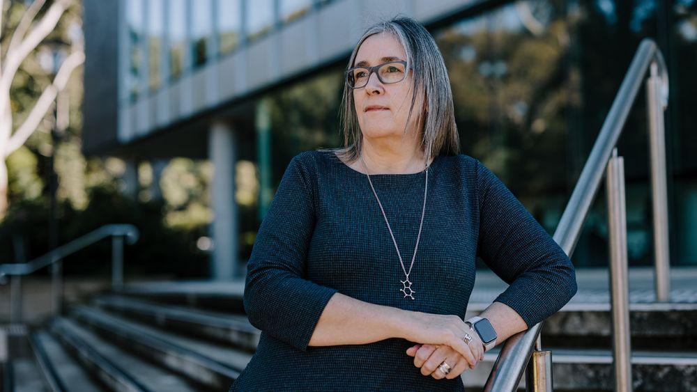 Jacqui Cameron stands thoughtfully near modern building stairs, wearing a dark dress, silver necklace, and smartwatch, in outdoor daylight.