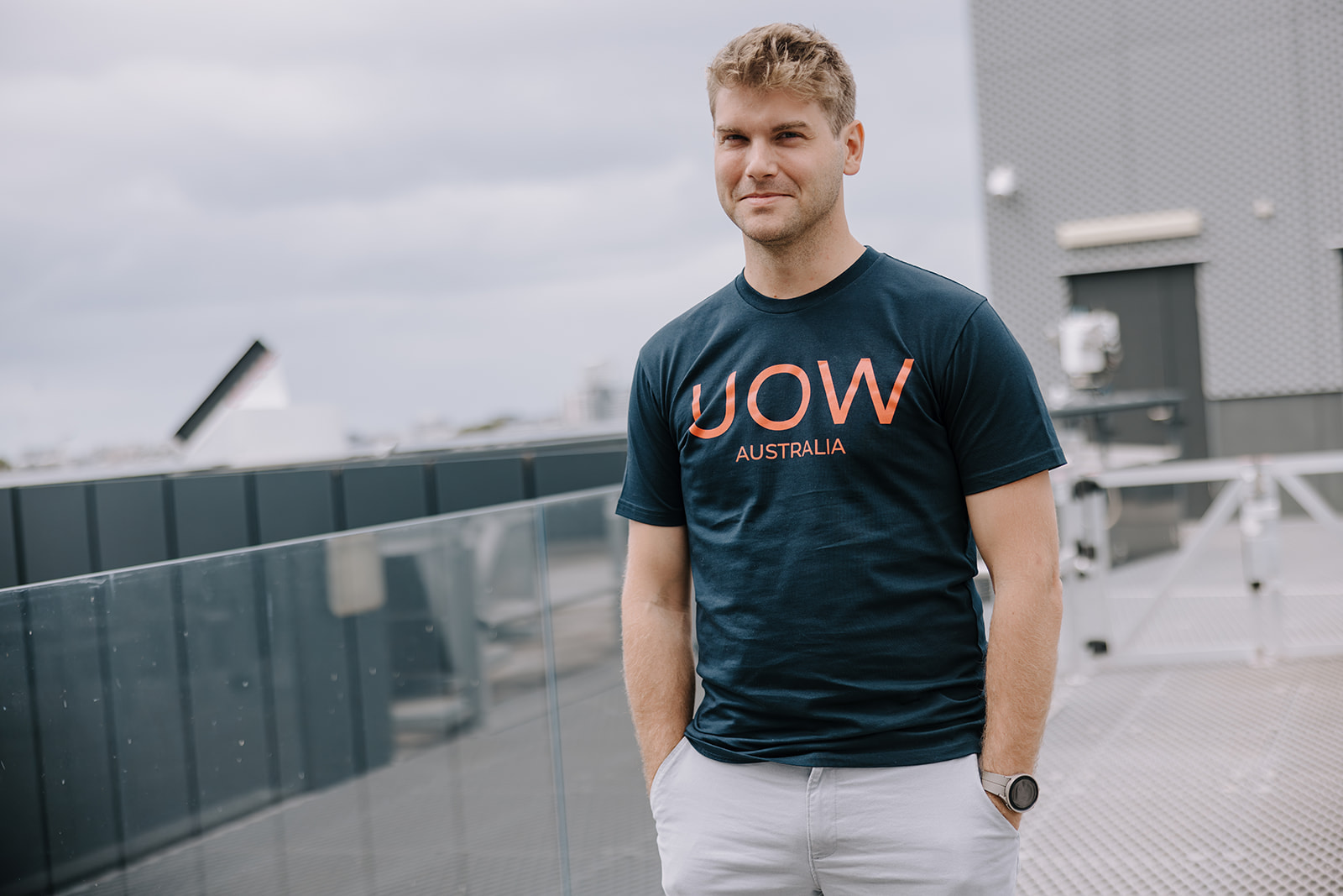 Max Desservettaz wears a navy UOW branded shirt and stands on top of a building with the sky in the background. Photo: Michael Gray