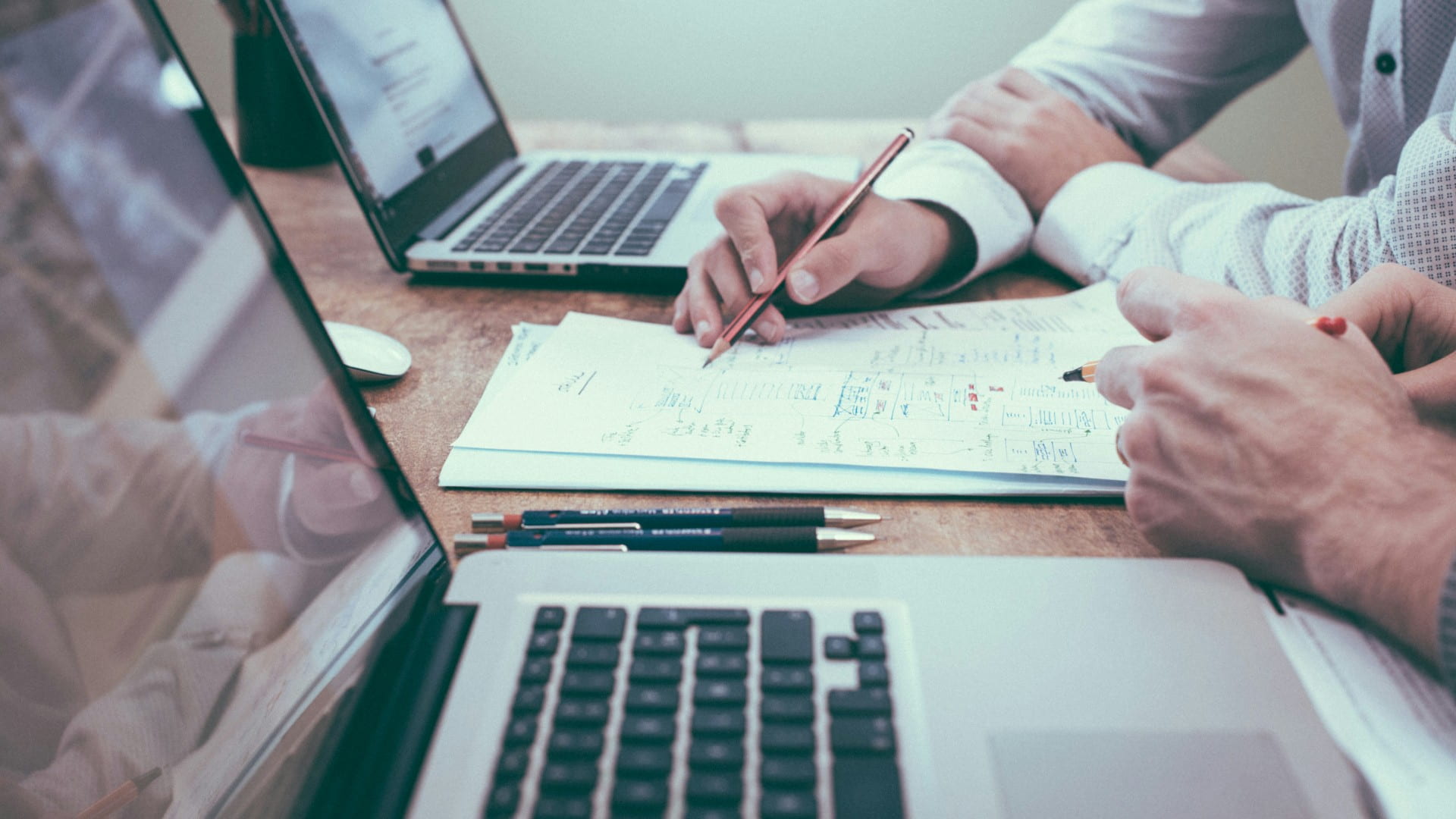 Two laptop computers are on a table with two people, only their forearms showing, are seen working. Photo: Scott Graham, Unsplash