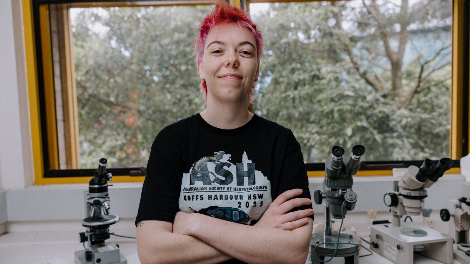 Researcher Kamryn Carter leans against a bench with her arms folded in a science lab, with two microscopes in the background. Photo: Michael Gray