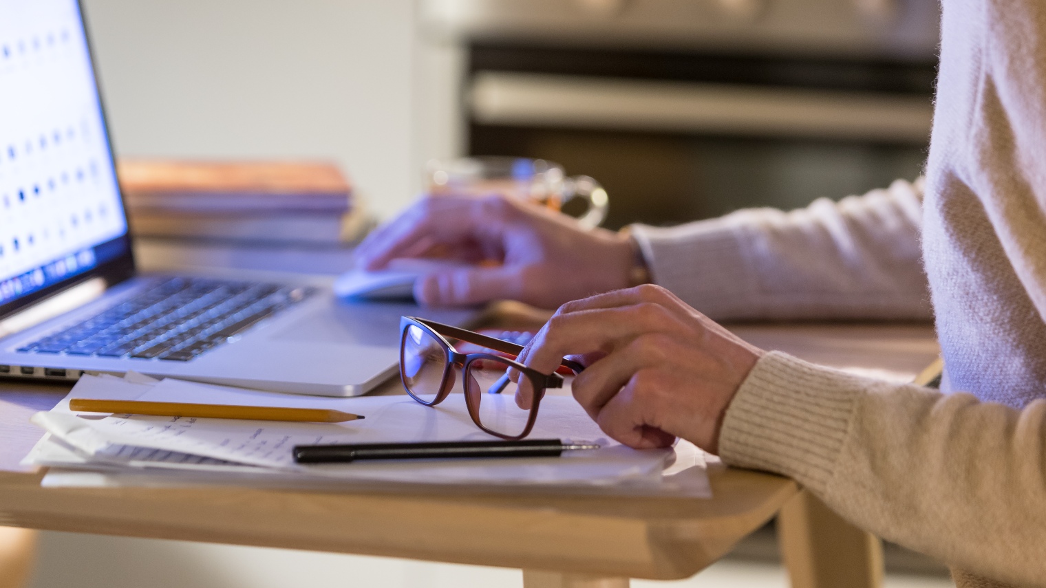 Closeup of man hands in beige sweater sitting and working at home at the laptop, holding eyeglasses, using computer mouse. Soft focus. Self isolation, stay home concept. Credit: Shutterstock Asset id: 1270782037