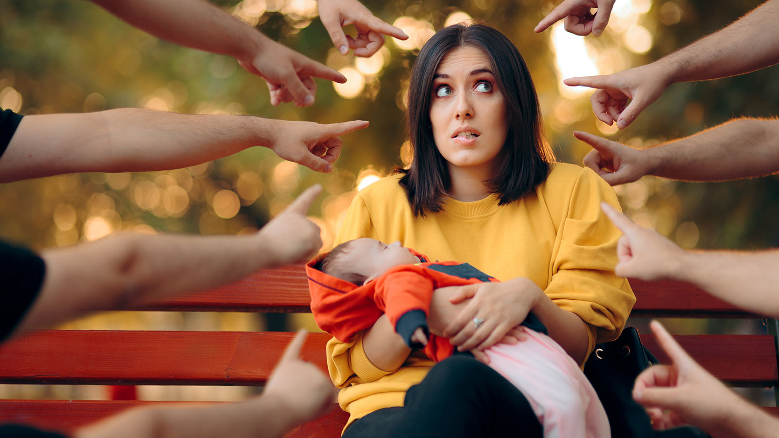 A large number of people point their fingers at a young mother sitting ion a bench with her baby