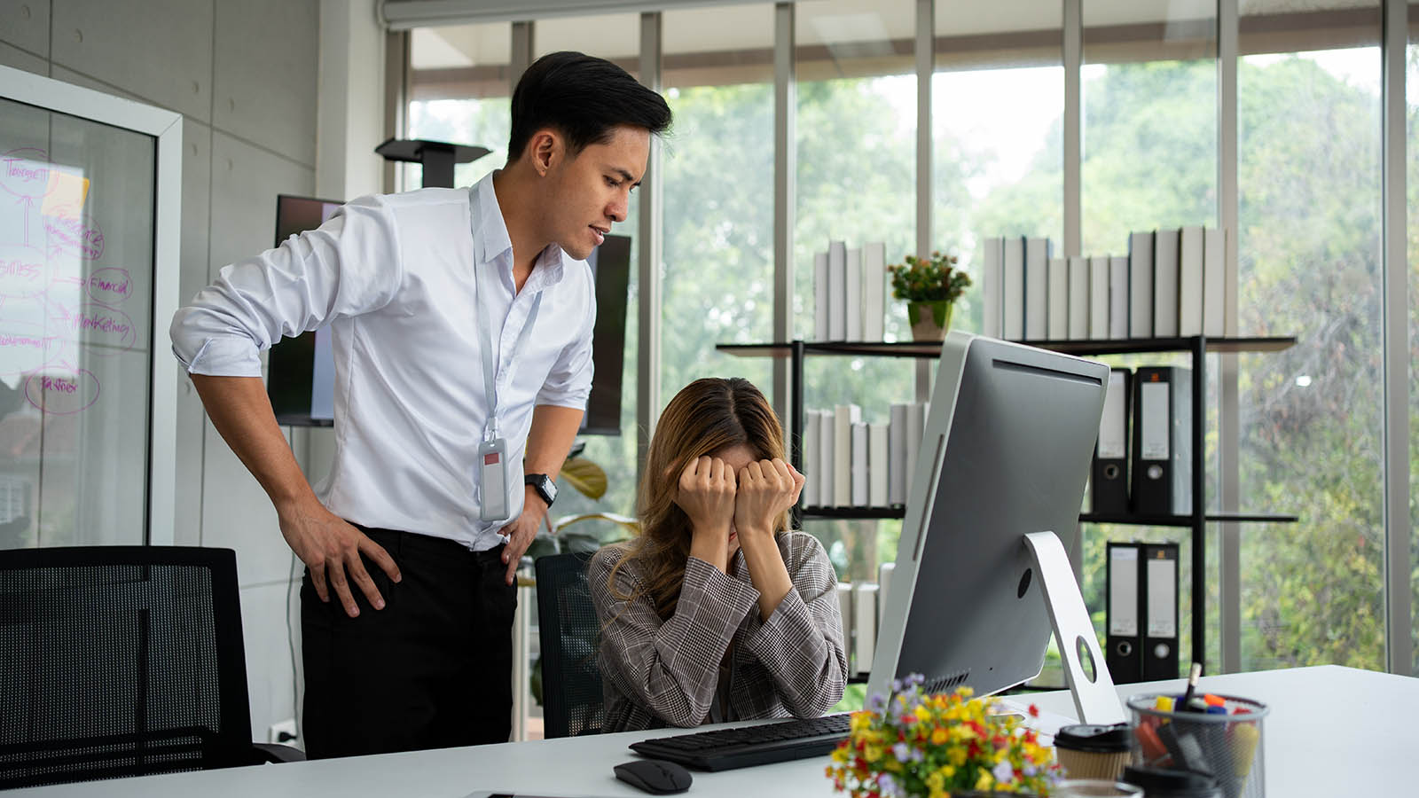 A stressed female office worker covers her face while a male colleague looks annoyed while looking at her computer screen.