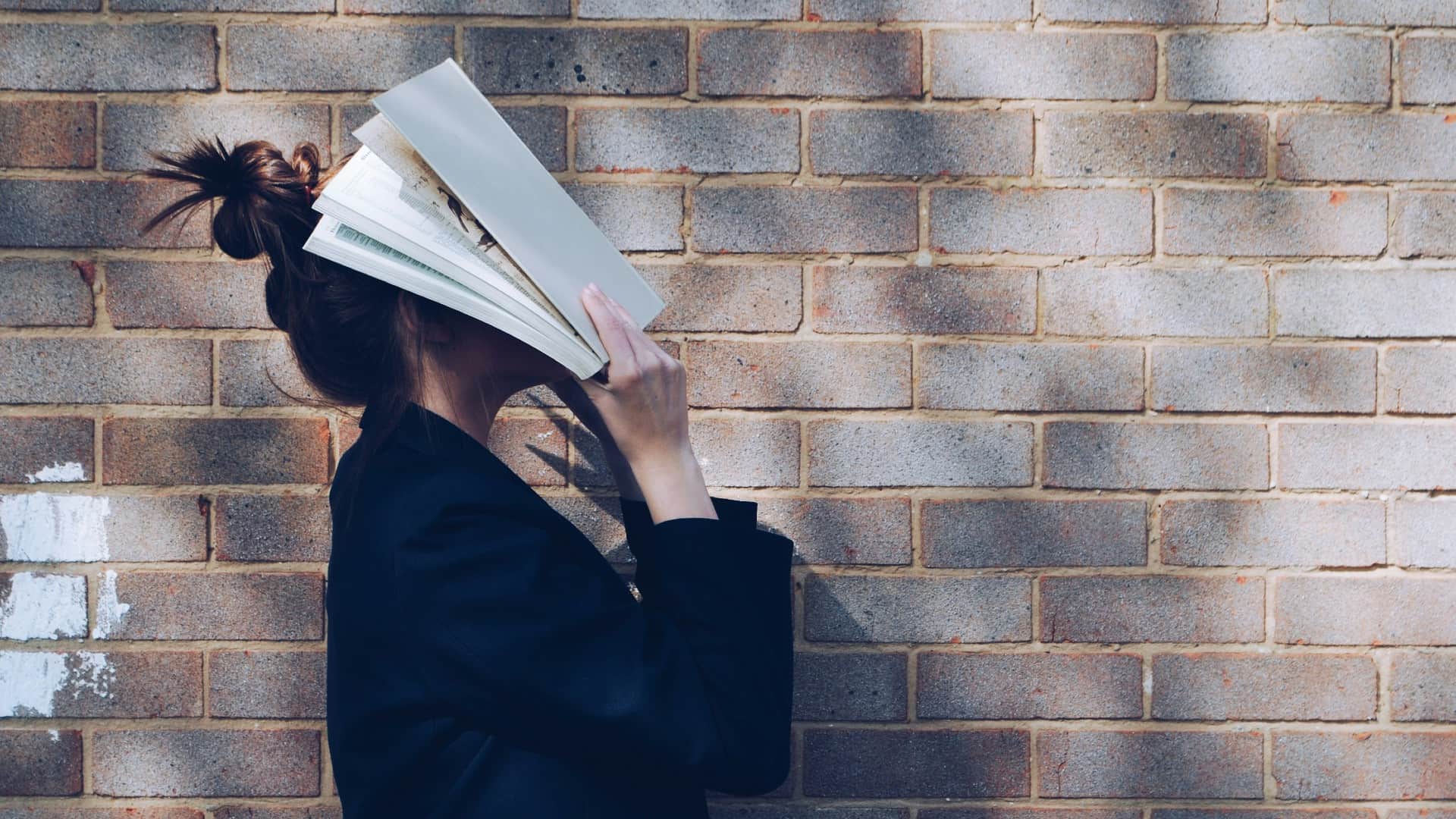 A women holds a book up to her face as she stands against a brick wall. Photo: Unplash