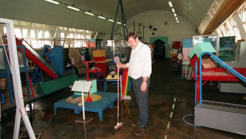 A man holding a broom in a large hall filled with various colorful exhibits and displays. The space has an arched ceiling and the floor appears slightly wet.